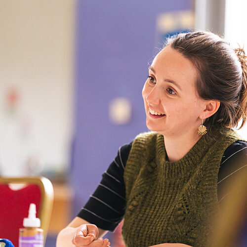 Smiling woman wearing a dark green jumper, sewing at a table