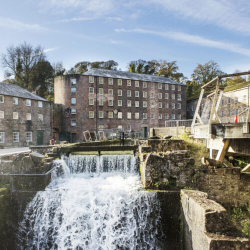 Water cascading down a tiered waterway with a bridge to one side in front of two large red brick mill buildings