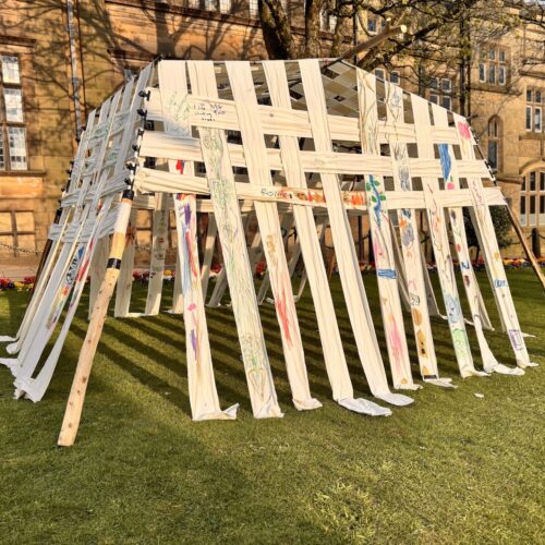 Temporary square structure overlaid in strips of fabric covered in colourful drawings, sited on grass outside an old building