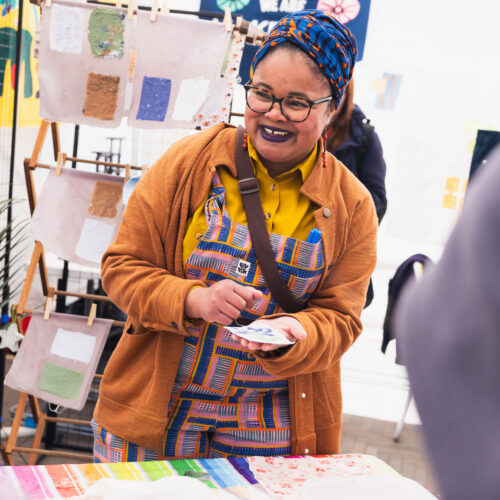 Photo of a black woman wearing a multi-coloured dress and turban and glasses, smiling and holding a textile square in her left hand. Behind her are bigger squares of painted textiles hanging on a wooden frame. She is standing inside a marquee next to a table with a multi-coloured tablecloth