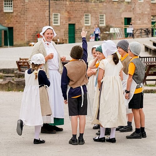 Woman in Victorian servant's clothing is surrounded by a group of children in Victorian clothing in front of a stone brick mill