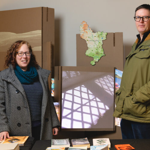 White woman wearing a grey coat with curly red hair and glasses stand in front of cardboard boxes and a table of books. At the other end of the table is a tall white man with dark hair and glasses and wearing a green parka
