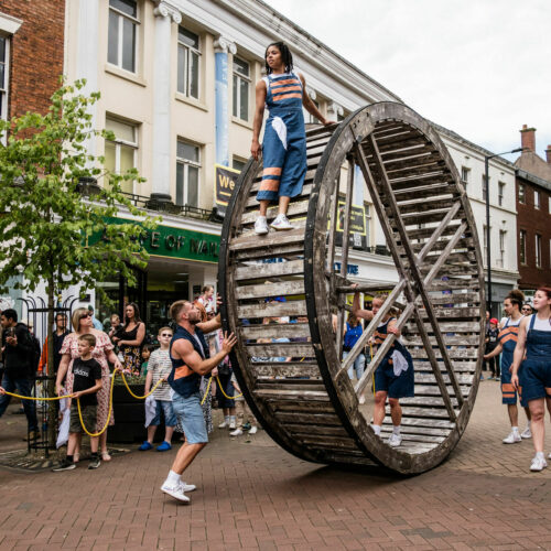 A street performer walks inside a large human-powered wheel while being guided along a town street, with a crowd watching the spectacle.