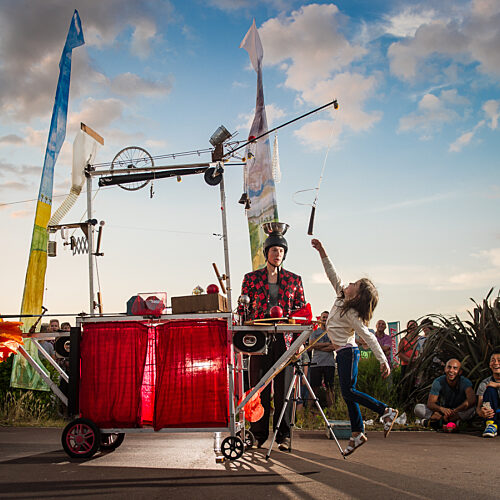 An outdoor street performance shows a performer operating a quirky mechanical contraption on wheels while another performer leaps beside it, with an audience gathered around watching.