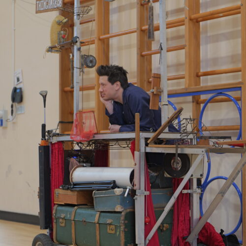 A performer leans thoughtfully on a large wheeled mechanical sculpture, resting his chin on one hand. The sculpture is assembled from vintage cases, metal frames, ropes and found objects inside a school hall.