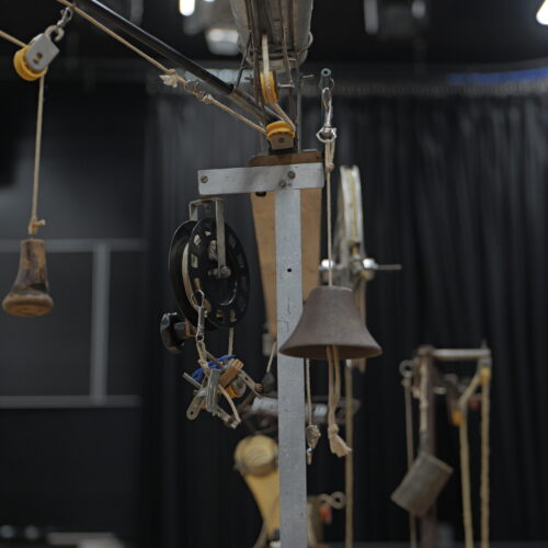 Close-up view of a kinetic sculpture featuring pulleys, ropes, bells and metal components suspended against a dark stage backdrop. The intricate arrangement highlights the handmade mechanical details.