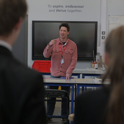 A workshop facilitator stands at the front of a classroom speaking to a group of students. The students are visible in the foreground, while a large screen and the words “To aspire, endeavour and thrive together” appear on the wall behind him.