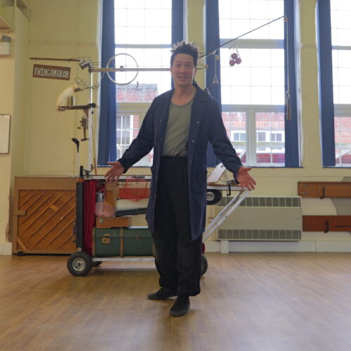 A performer stands in a school hall with arms open in welcome beside a large wheeled mechanical sculpture made from metal frames, pulleys, bells and found objects. Tall windows fill the space with natural light.