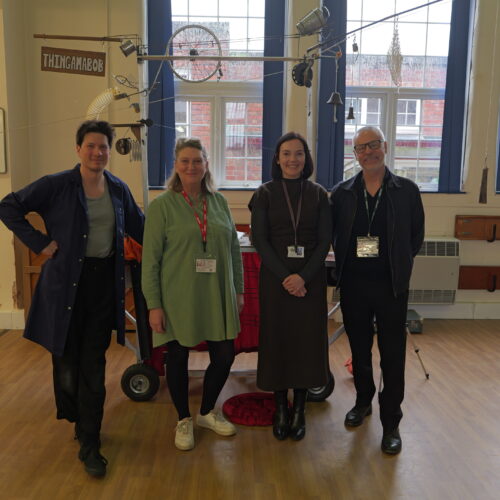 Four adults stand together in a school hall in front of a large kinetic sculpture. They smile towards the camera, with the sculpture’s framework and hanging objects visible behind them.