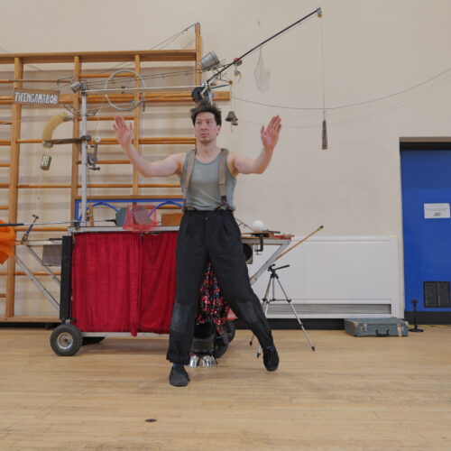 A performer poses in front of a kinetic sculpture in a school hall, arms raised as if conducting or controlling the machine. The sculpture, made from pulleys, ropes and assorted objects, stands behind him against a gym wall.