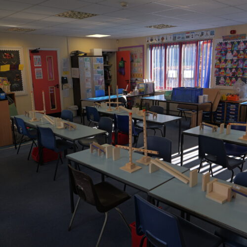 Empty classroom set up for a hands-on engineering or design activity, with wooden construction kits arranged on tables between blue chairs. Sunlight streams through tall windows, and colourful classroom displays decorate the walls.