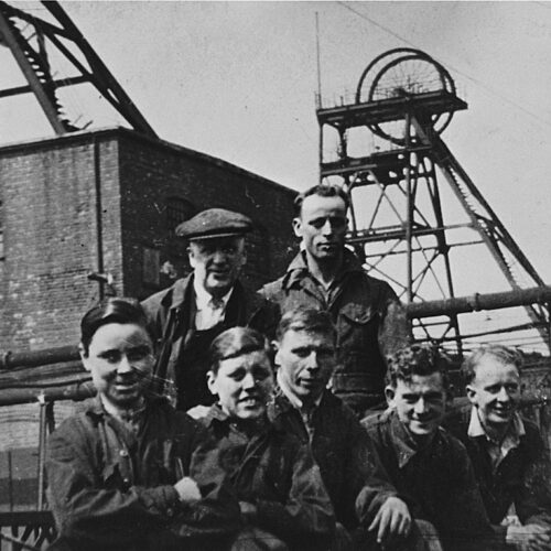 Black and white photo of miners stood in front of a Pit Head in a colliery