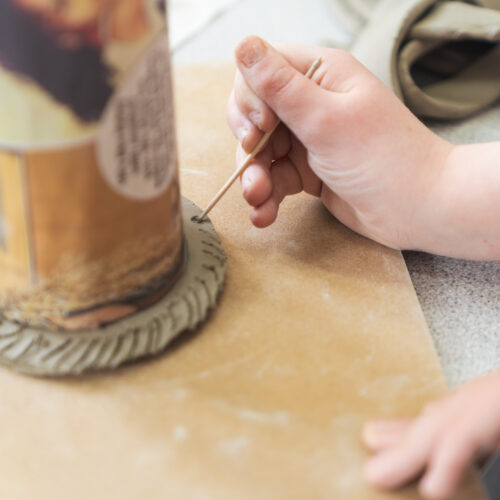 A hand uses a tool to carve details into soft clay around a cylindrical object.