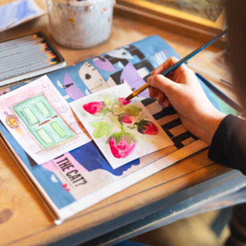 A person paints a small artwork with a brush, with coloured pencils and materials laid out nearby.