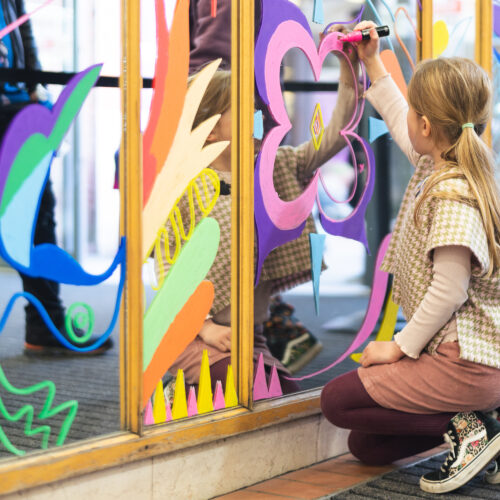 A young child kneels and draws bright shapes on a glass window as part of a creative activity.