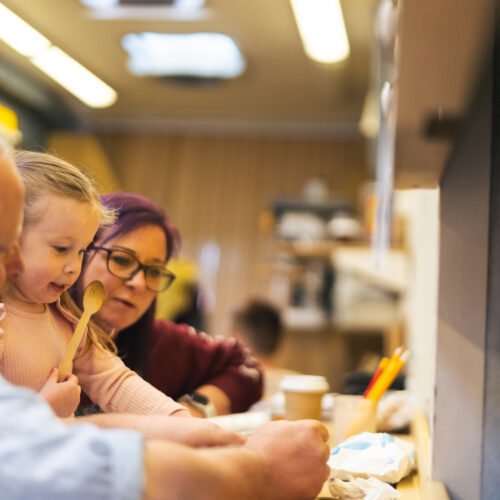 An adult and child work together at a table, with the child holding a wooden spoon during a hands-on activity.