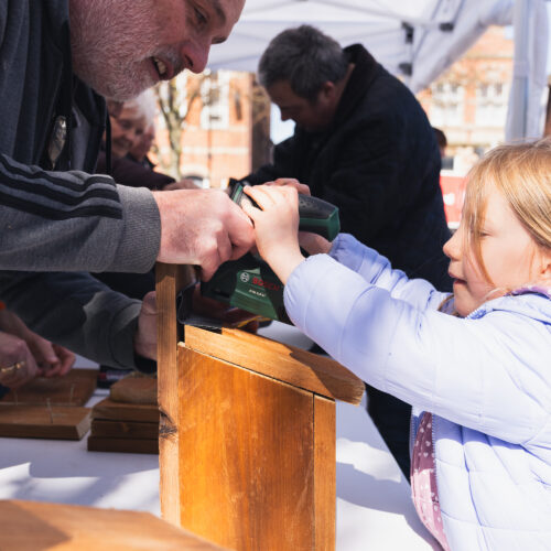 A child uses a power tool with guidance from an adult to build a wooden structure at an outdoor workshop.
