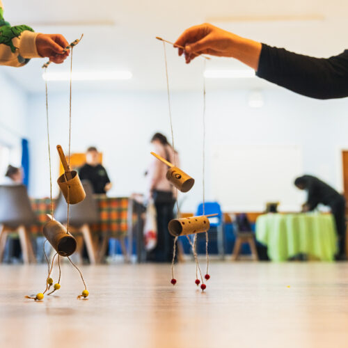 Two people hold simple handmade puppets on strings, experimenting with movement in a workshop space.