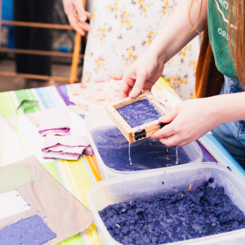 Hands hold a small frame dripping with pulp over a tray, demonstrating a papermaking process.