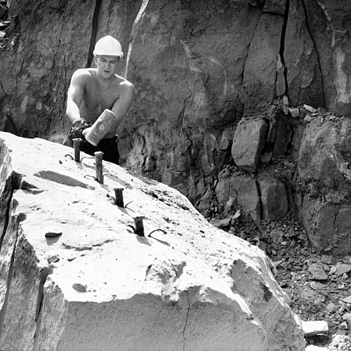 Black and white photo of a bare-chested quarryman hammering metal plugs in a piece of stone in a quarry