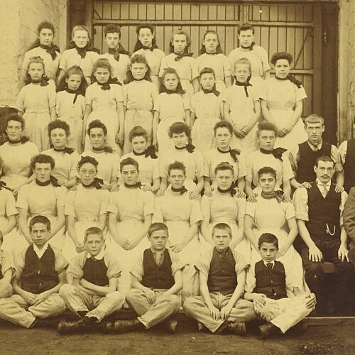 Cepia photo of lines of seated girls and young women wearing white dresses and dark neckerchiefs, a row of boys seated at the front wearing dark waistcoats and light trousers, plus three men sat at the side wearing dark waistcoats and trousers, and an old man with whiskers wearing a woollen suit, holding a hat